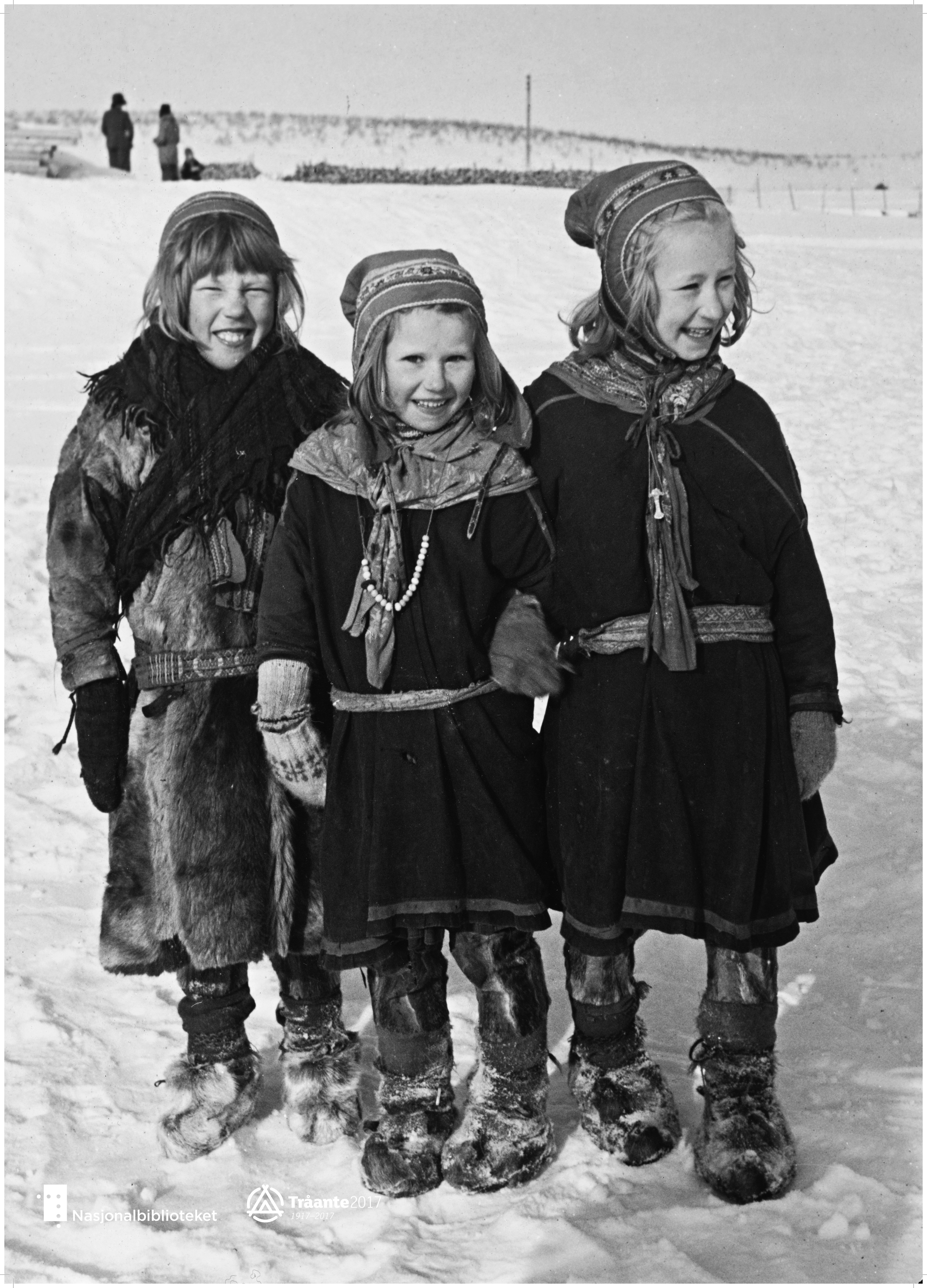 Tre jenter i kofter som smiler til kamera. Snø, og sort hvitt.