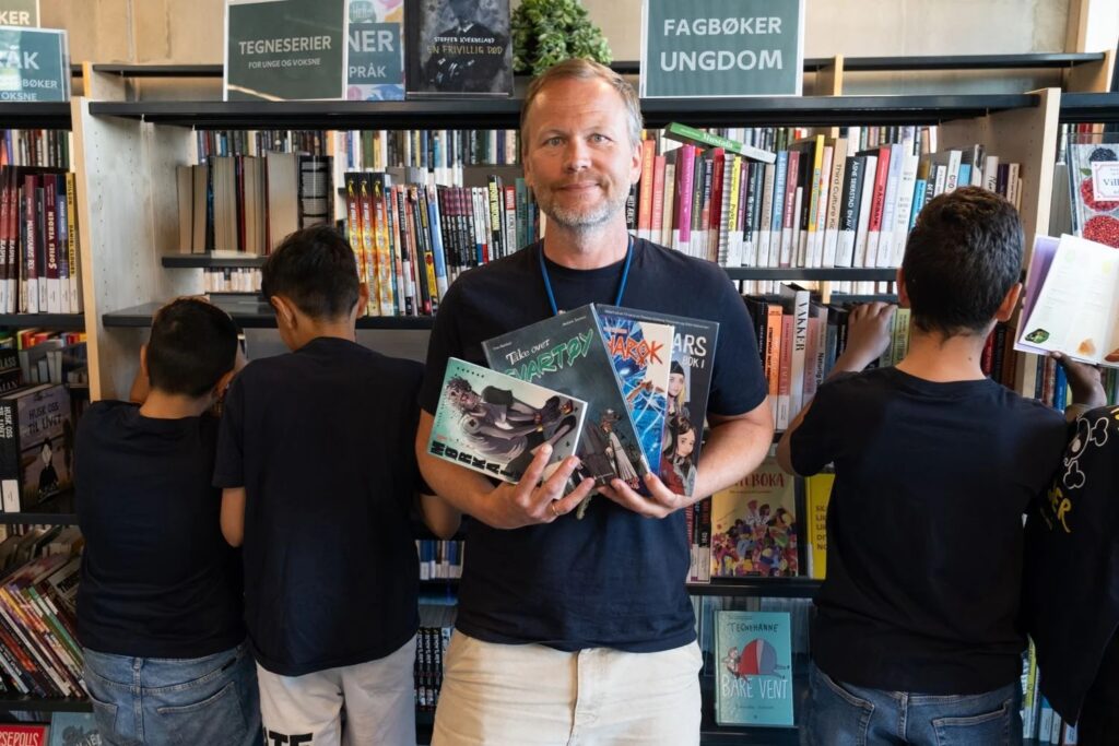 Jørgen Gromstad holder opp bøker foran bokhylle i biblioteket