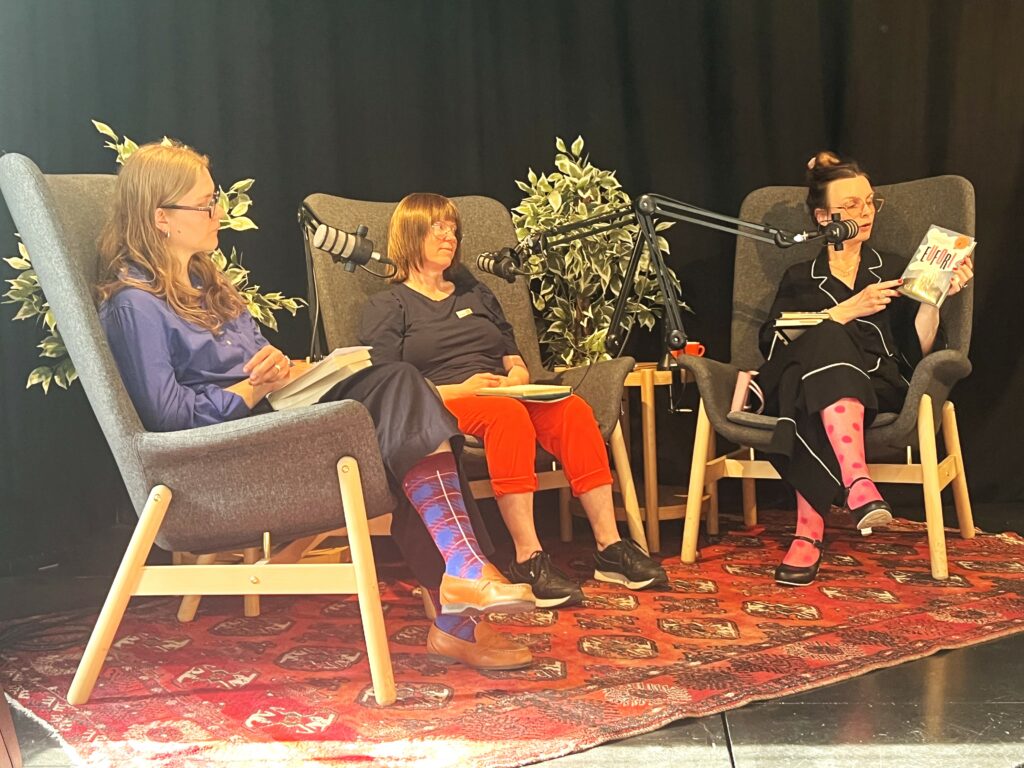 Tre kvinner sitter i stoler på en scene med mikrofoner. En kvinne viser fram en bok