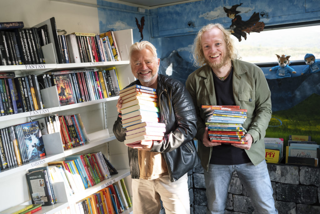 To menn står med bøker i henda foran ei bokhylle med bøker
