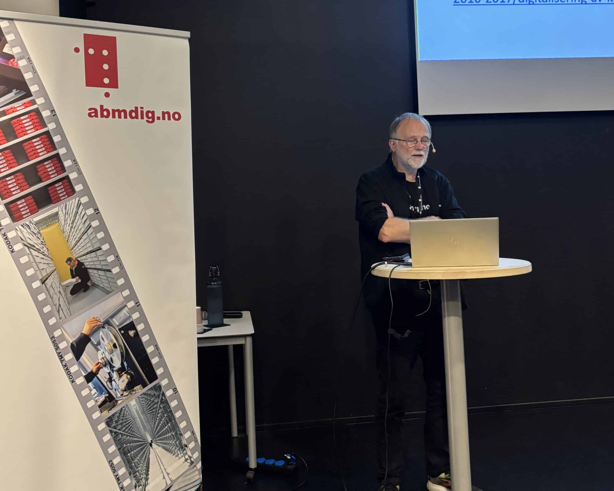 Foredragsholder står ved siden av roll-up med bilder og logo for abmdig.no