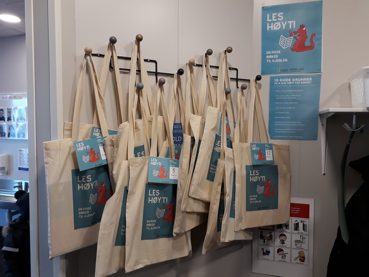 Flere titals tøyvesker henger på knaggrekke i garderoben, og plakat med informasjon om tøyveskebiblioteket og gode grunner til høytlesning er satt opp like ved.