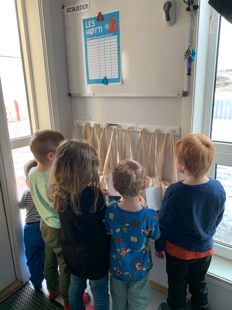 Fem barn unedrsøker tøyveskebiblioteket og utlånsplakat for å skrive seg opp som låner henger på veggen.