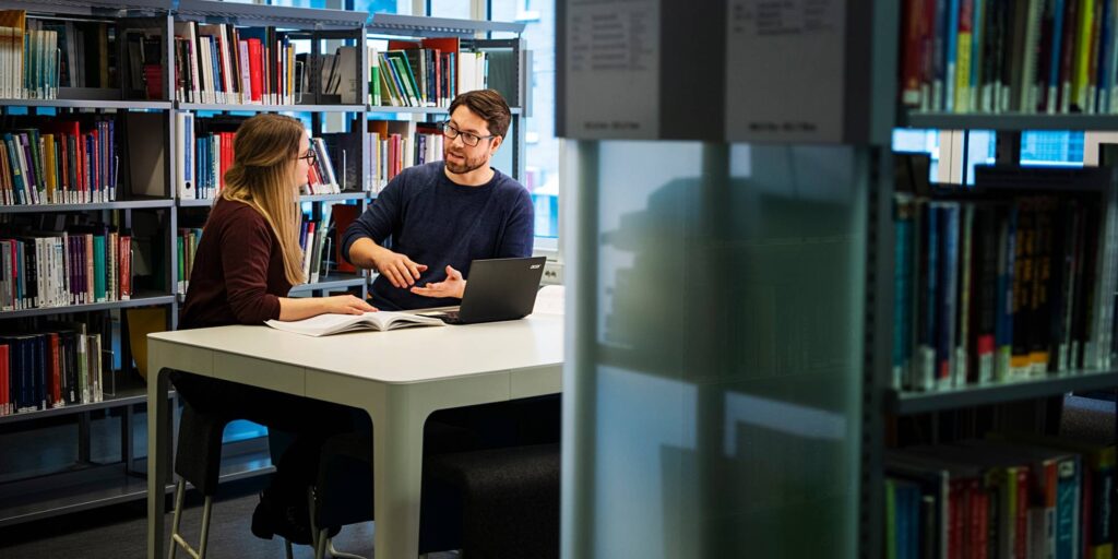 To personer sitter ved et bord mellom hyller i et fagbibliotek